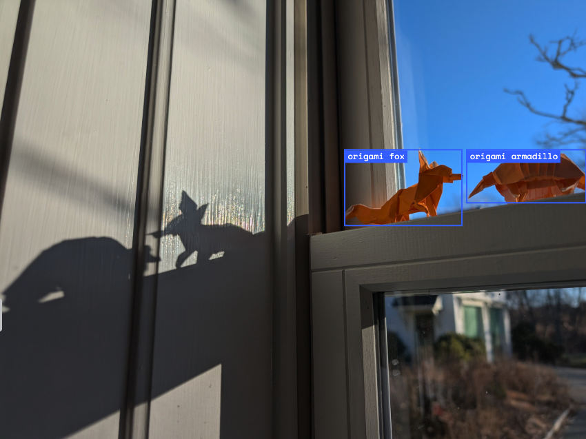 Origami fox and armadillo on a windowsill, casting shadows on a sunlit wall, with a clear blue sky visible outside. 
